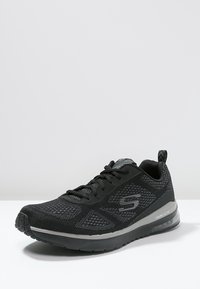 Černá sportovní obuv Skechers s vrchní částí z prodyšné síťoviny, polstrovanou podešví a poutkem na patě, umístěná na bílé ploše s jednoduchým šedým pozadím.