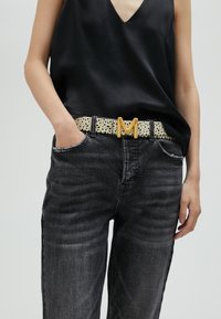 Top en satin noir, jean gris taille haute, et une ceinture crème avec des pois noirs et une boucle en or en forme de "M". Tissu texturé et accents distincts.