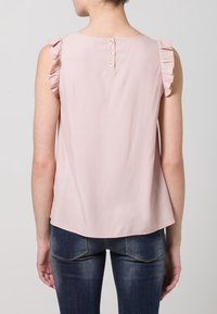 Světle růžový top bez rukávů s volánkovými akcenty na ramenech, zapínání na knoflíky na zádech a hladkou texturou. Kombinováno s tmavě modrými džíny.