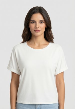 Femme aux cheveux bruns mi-longs portant un t-shirt blanc à manches courtes uni et un jean bleu clair, debout devant un fond uni.
