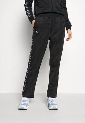 Personne portant un pantalon de jogging noir avec des bandes logo sur le côté et des baskets bleu clair et blanches, debout sur un fond uni.