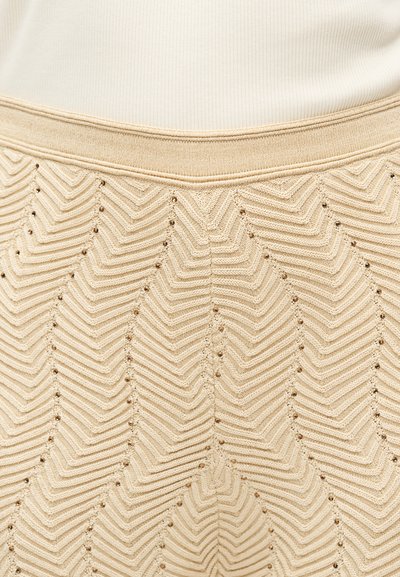 Μπεζ πλεκτή φούστα με σχέδιο ζικ-ζακ και μικρές οπές, φορεμένη με ριμπ κρεμ τοπ που είναι μπουκωμένο στη μέση της φούστας.
