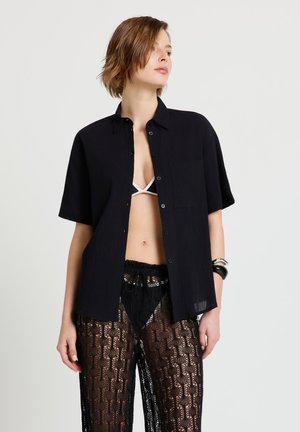 Jeune femme portant une chemise noire à manches courtes ouverte sur un haut de bikini blanc et un pantalon en dentelle noire transparente avec des bracelets au poignet.