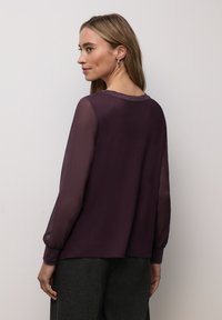 Top púrpura con mangas largas transparentes, cuello redondeado y puños elásticos. Tela suave con un brillo sutil, combinada con pantalones gris oscuro.