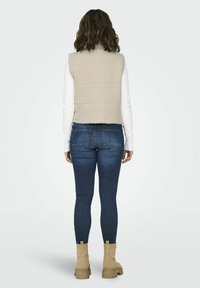 Gilet en polaire beige avec un col haut, des plis au dos et un design court, porté sur une chemise blanche à manches longues. Associé à un jean skinny bleu marine et des bottes beiges.