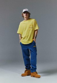 T-shirt en coton jaune avec poche et logo, associé à un pantalon bleu avec logo, et des bottes marron à lacets sur fond gris.