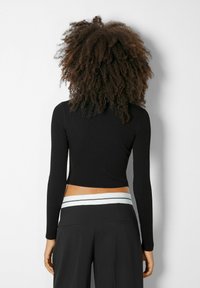 Černý žebrovaný crop top s dlouhými rukávy a přiléhavým střihem, zobrazený zezadu, doplněný černými kalhotami s kontrastním pasem.