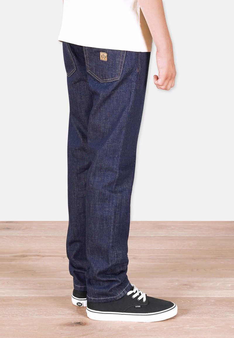 Tummansiniset farkut suorilla lahkeilla, joissa on kontrastiväriset tikkaus ja pieni logo-ompelu takataskussa. Mustat tennarit valkoisilla nauhoilla.