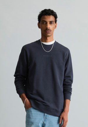 Jonge man met krullend haar die een donkerblauwe sweatshirt draagt, lichtblauwe spijkerbroek, een zilveren ketting en armband, staand met één hand in de zak.