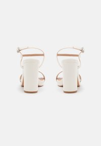 Sandales à talons blancs avec un talon carré, en matériau brillant, à l'arrière ouvert, et de fines brides autour de la cheville avec un détail de boucle décoratif.