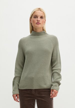 Blonde vrouw draagt een lichtgroene gebreide trui met lange mouwen en een hoogsluitende kraag, gecombineerd met donkerbruine broek.