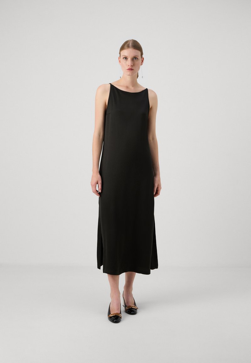 Rochie neagră fără mâneci, cu o siluetă fluidă, având o textură netedă și fante laterale. Asortată cu pantofi negri și accesorii simple.