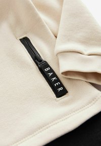 Sudadera beige con tejido texturizado. Presenta un bolsillo con cremallera negro y una etiqueta negra de marca con letras blancas en el lateral.