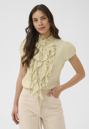 Kobieta z długimi ciemnymi włosami, ubrana w jasnożółtą bluzkę z falbanami i jasnobeżowe spodnie z wysokim stanem, pozująca z jedną ręką w kieszeni.