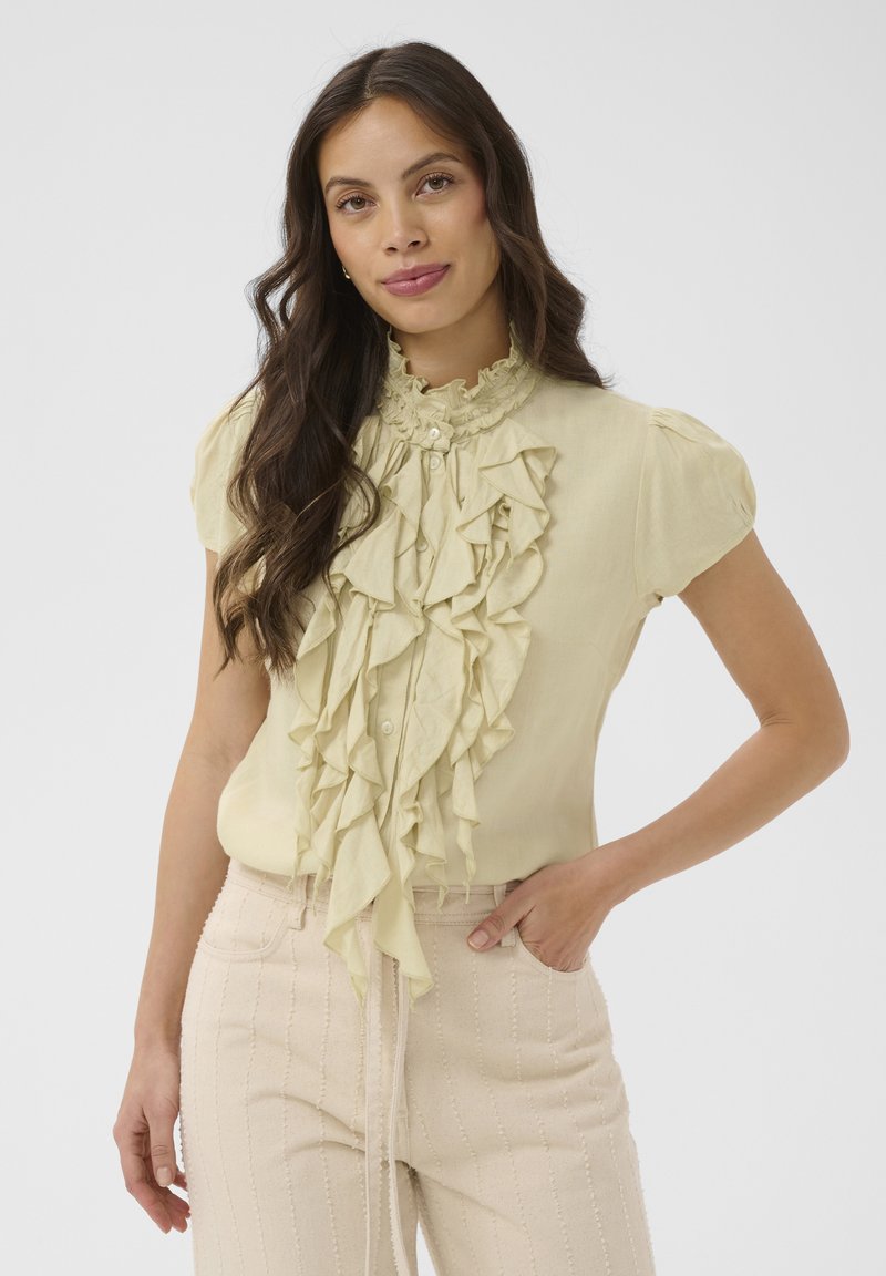 Femme aux longs cheveux noirs portant un chemisier jaune pâle à volants et un pantalon taille haute beige clair, posant avec une main dans la poche.