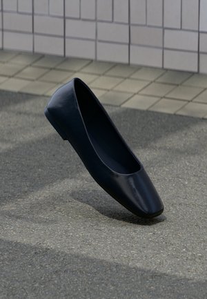 Zapato plano de cuero negro, de pie verticalmente sobre una superficie de asfalto cerca del borde de una acera con baldosas.