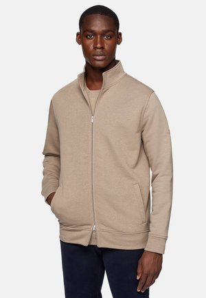 Felpa beige con zip e colletto alto, tasche laterali, polsini a costine e tessuto strutturato. Indossata sopra un top coordinato.