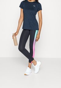 Kvinna i marinblå träningströja, svarta leggings med gradient i rosa och teal sidan, vita sneakers, hållande en beige vattenflaska.