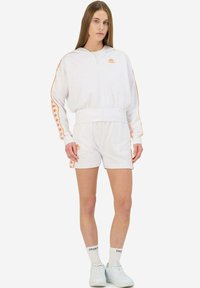 Sudadera blanca con cremallera y acentos naranjas, combinada con pantalones cortos blancos. Cuenta con un logo en el pecho y rayas laterales. Modelo lleva zapatillas blancas.