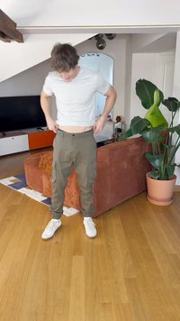 Pantaloni cargo verdi oliva con tasche laterali e polsini elasticizzati, abbinati a una camicia bianca a righe e sneakers bianche, su un pavimento in legno.
