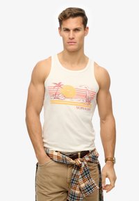 Top sin mangas blanco con un gráfico colorido que incluye un sol, palmeras y patrones a rayas. Combinado con pantalones beige y una camisa de cuadros atada alrededor de la cintura.