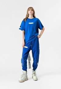 Camiseta azul oversize con un detalle de etiqueta blanca, combinada con joggers azules que tienen bordado de plumas y un bolsillo lateral, llevada con botas blancas.