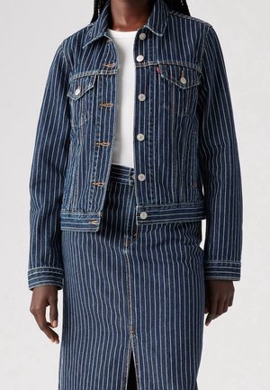 Mørkhudet person iført en blå denimjakke med nålestriber og matchende nederdel med en hvid top under, stående mod en ensfarvet baggrund.