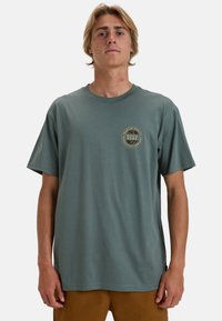 Baumwoll-T-Shirt in gedämpftem Grün mit kurzen �Ärmeln, das ein kreisförmiges Logo auf der linken Brust in Beige und Schwarz zeigt. Lässige Passform, Rundhalsausschnitt.