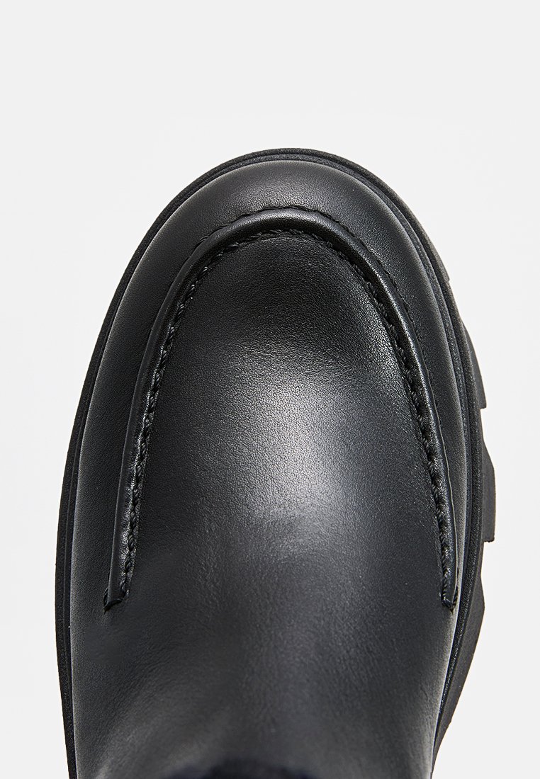 Schwarzer Lederschuh mit abgerundeter Spitze, sichtbar genäht und mit einer strukturierten Oberfläche. Die Sohle ist dick und leicht rugged.