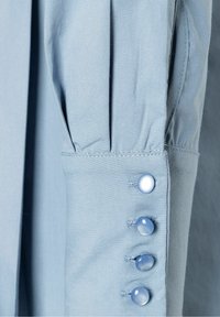 Blusa azzurro chiaro con maniche a pieghe e quattro bottoni abbinati con finitura lucida, che evidenziano la texture del tessuto morbido e le cuciture pulite.