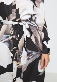 Nahaufnahme eines gebuttonierten Blumenkleidungsstücks mit einem gebundenen Taillengürtel und einer sichtbar am Körper ruhenden Hand, das Schwarz, Weiß, Beige und Blautöne aufweist.