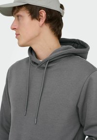 Jeune homme portant un sweat à capuche gris et une casquette beige, regardant vers la gauche sur un fond clair uni.