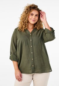 Chemise olive, à boutons, avec un col en V, des manches trois-quarts et des détails décoratifs à rouleau. Fabriquée en tissu léger et texturé.