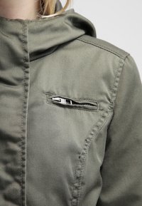 Olivově zelená bunda s kapucí, předním zipem a zipovou náprsní kapsou. Vyrobena z měkké látky s viditelnými švy.