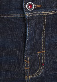 Vaqueros de mezclilla azul oscuro con un diseño tradicional de cinco bolsillos, costuras de cobre y un botón metálico con un detalle en rojo.