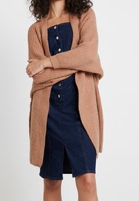 Žebrovaný, měkký, béžový cardigan přeložený přes přiléhavé, tmavé džíny šaty s předními knoflíky a rozparkem, vytvářející kontrast barev a textur.