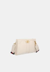 Beige leather handbag with a gold clasp, structured design, detachable strap, and a small attached tag with a multi-colored accent.