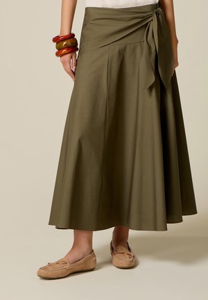 Olijfgroene midi-rok met taillekoord, gedragen met beige mocassins en dikke amberkleurige armbanden aan de linkerpols.