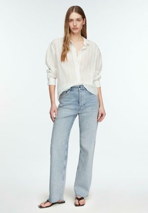 Junge Frau mit langen Haaren, die ein lockeres weißes Hemd mit Knöpfen, hellblaue hochtaillierte Jeans und schwarze Flip-Flops trägt und vor einem weißen Hintergrund steht.