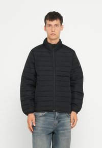 Jachetă neagră padded cu guler înalt, fermoar pe toată lungimea frontală și manșete elastice. Prezintă cusături orizontale și un design minimalist.