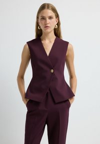 Gilet senza maniche a scollo a V in tessuto bordeaux scuro, con un bottone dorato, silhouette sartoriale e orlo a peplum. Texture liscia, design minimal.
