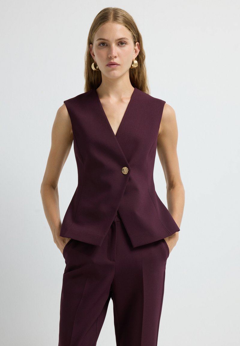 Gilet senza maniche a scollo a V in tessuto bordeaux scuro, con un bottone dorato, silhouette sartoriale e orlo a peplum. Texture liscia, design minimal.