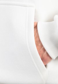 Mano dentro della tasca anteriore di una felpa bianca con polsino a coste visibile sul polso.