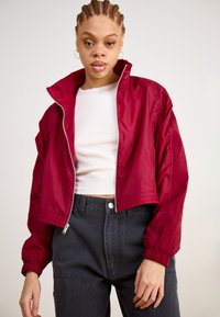 Red windbreaker jacket with a high collar, zip front, and elastic cuffs, paired with a white cropped top and dark gray high-waisted pants.