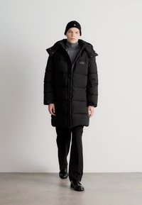 Black puffer coat with a hood, featuring a front zipper, quilted texture, and a brand label on the upper left side. Model wears black pants and shoes.