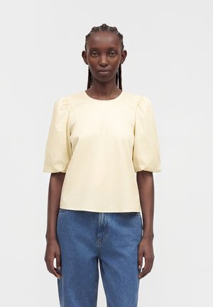 Femme aux cheveux tressés portant un chemisier jaune pâle à manches bouffantes et un jean bleu, debout devant un fond blanc uni.