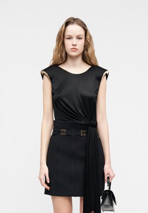 Kobieta o długich falowanych włosach nosi czarną sukienkę bez rękawów z detalem węzła i paskiem, trzymając małą czarną torebkę na jednolitym tle.