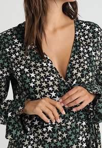 Sukienka typu wrap z czarnym tłem i zielonymi, białymi oraz beżowymi wzorami w gwiazdki. Dekolt w serek, długie rękawy oraz miękka tekstura materiału.