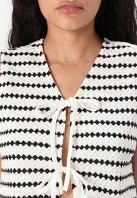 Strikket crop top med et sort og hvidt zigzag mønster, V-udskæring og en sløjfe lukning foran. Blød tekstur.