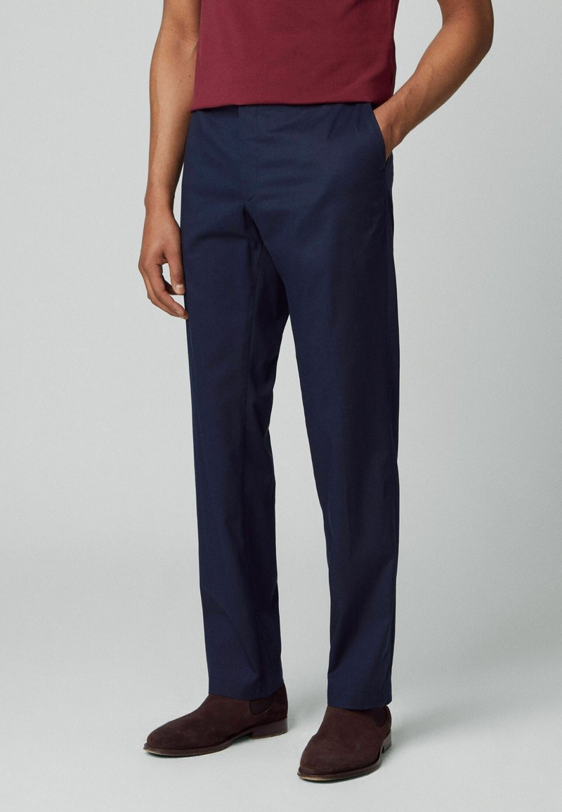 Homme portant un pantalon de costume bleu marine et des chaussures en daim marron foncé, une main dans la poche et une chemise bordeaux visible.